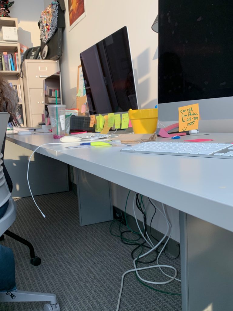 Two computers with many sticky note reminders attached to them sit on a desk in an office.